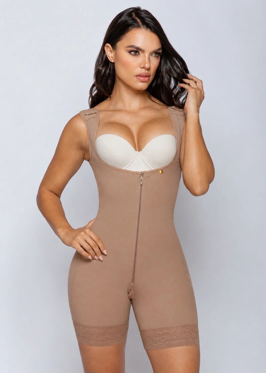 A woman modeling a beige full-body compression shapewear bodysuit with a front zipper, built-in underwire bra cups, and lace trim at the thighs, against a light gray studio background.
