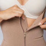 Close-up of a person wearing a beige shapewear bodysuit with a front zipper, lifting the side fabric with both hands to reveal a white underwire bra underneath.
