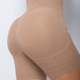 Close-up of a model wearing high-waisted beige shapewear shorts with a wide elastic leg band, shown from the back against a light gray background.