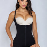 A woman modeling a black shapewear bodysuit with a white underwire bra insert, front zipper, and lace leg shorts against a light gray studio background.