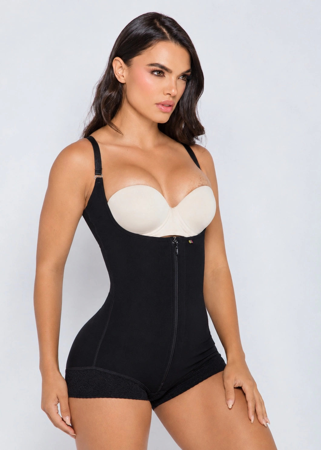 A woman modeling a black shapewear bodysuit with a front zipper and a contrasting beige underwire bra insert, set against a plain light gray studio background.