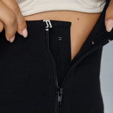 Close-up of a person wearing black shapewear or leggings with a front zipper, pulling the fabric outward with both hands to reveal the interior lining. A cream bra is visible at the top, and the person's midriff and hands are shown against a plain light gray background.