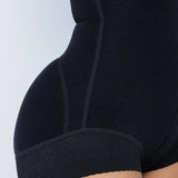 Close-up of a person wearing black shapewear briefs, showing the side hip, waist seam, and textured leg band against a light gray background.