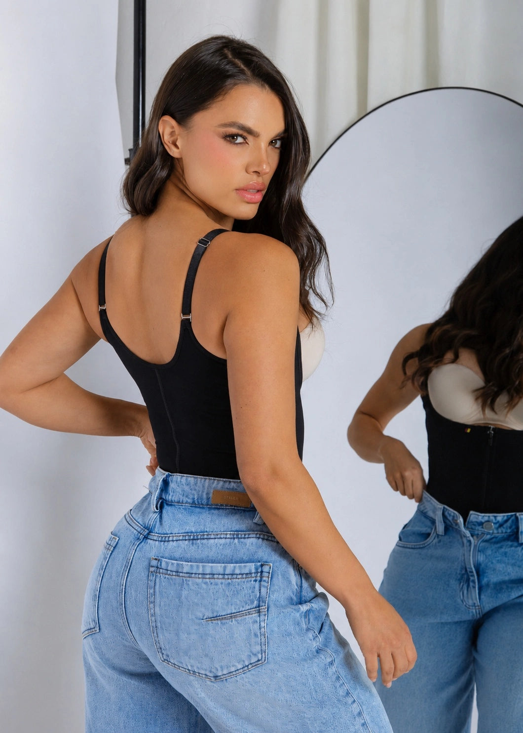 A woman posing in a black strappy corset-style camisole top and light-wash blue jeans, with her reflection visible in a studio mirror.