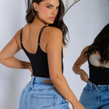 A woman posing in a black strappy corset-style camisole top and light-wash blue jeans, with her reflection visible in a studio mirror.