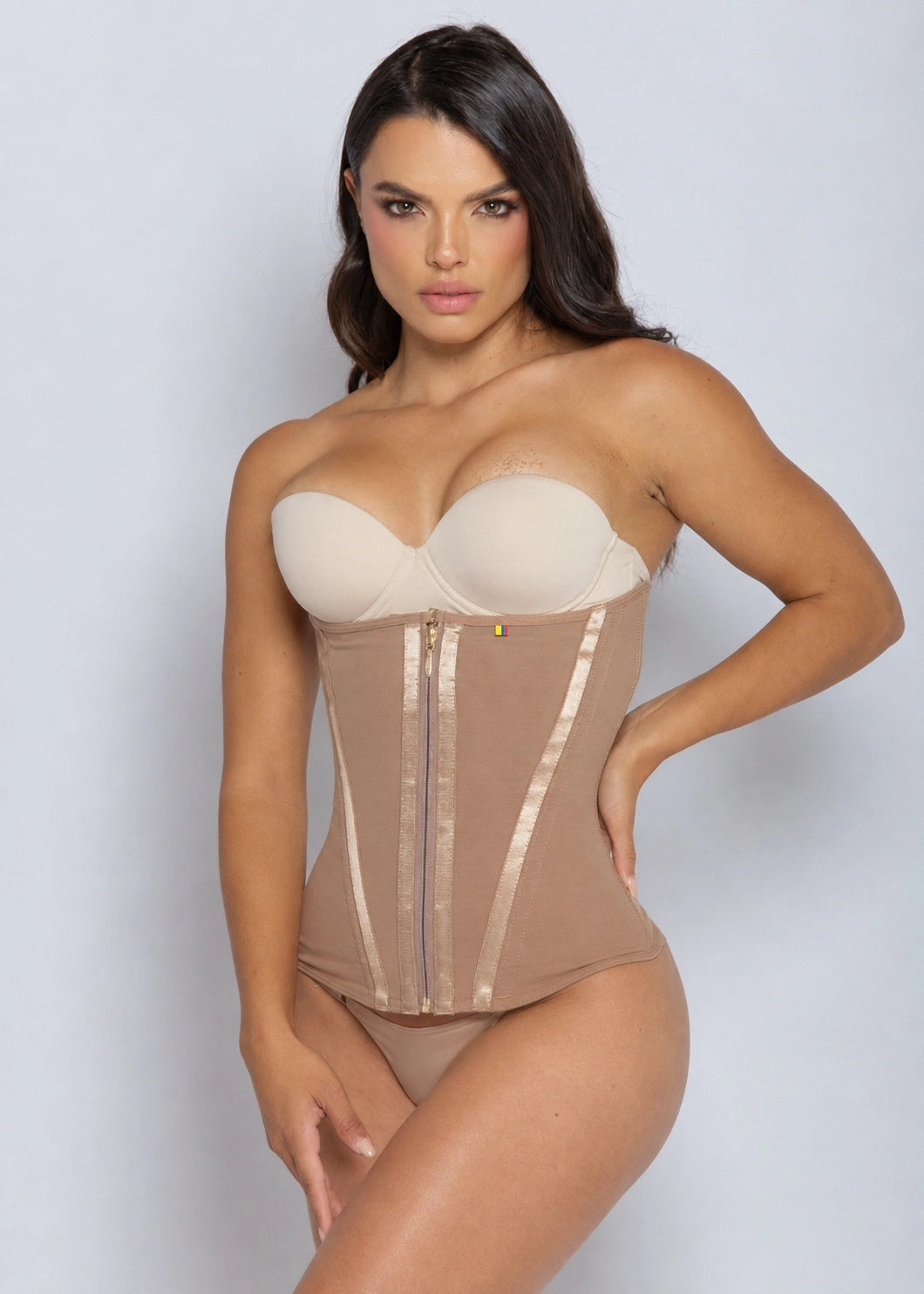 A woman modeling a strapless beige corset bodysuit with a structured bust, sheer mesh panels, vertical satin-like trim, and a front zipper, against a light gray studio background.
