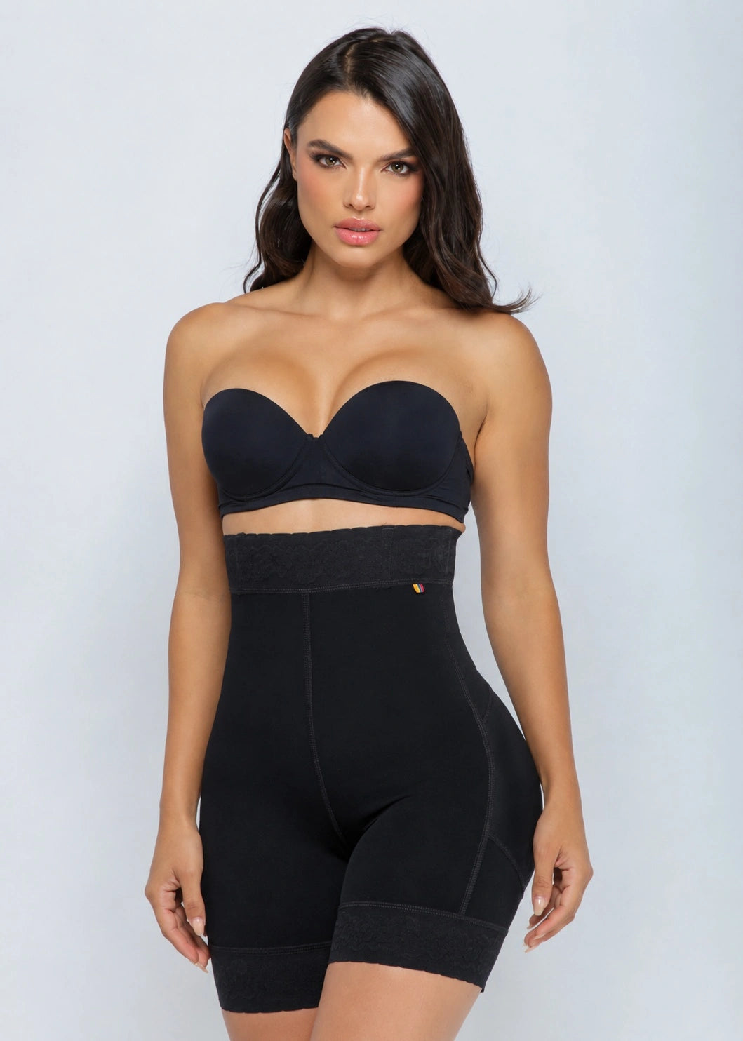 A woman modeling a black strapless bandeau-style top and high-waisted shapewear shorts set against a light gray studio background.