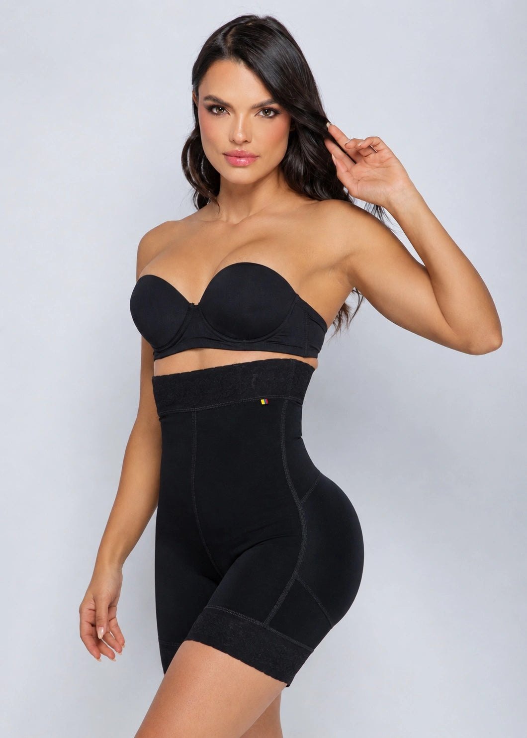 A woman modeling a black shapewear set consisting of a strapless bandeau-style bra and high-waisted briefs, photographed against a light gray studio background.