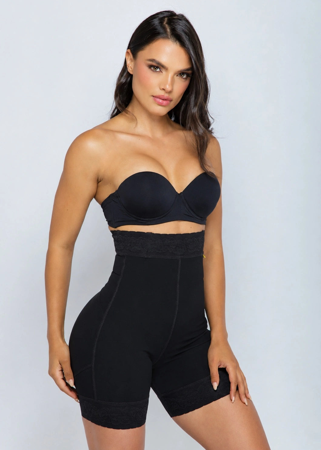 A woman modeling a black strapless bandeau bra and matching high-waisted black shapewear shorts against a light gray studio background.