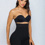 A woman modeling a black strapless bandeau bra and matching high-waisted black shapewear shorts against a light gray studio background.