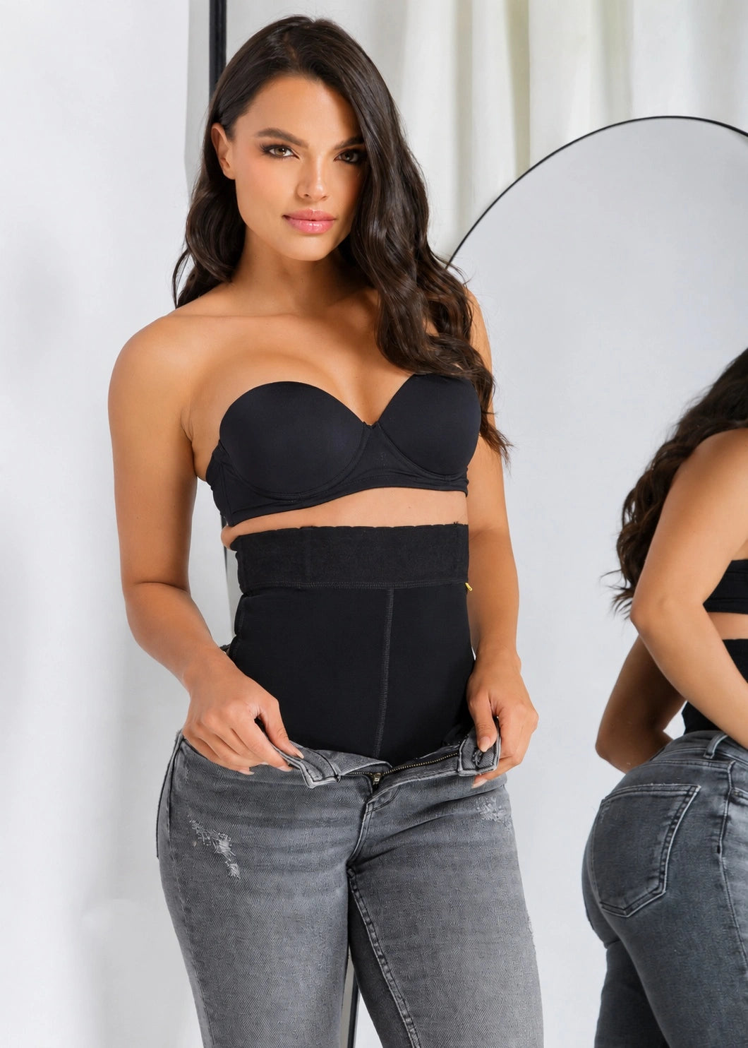 A woman modeling a black strapless bandeau-style top and matching high-waisted black waist trainer or shapewear, paired with gray jeans, with a mirror visible in the background.