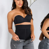A woman modeling a black strapless bandeau-style top and matching high-waisted black waist trainer or shapewear, paired with gray jeans, with a mirror visible in the background.
