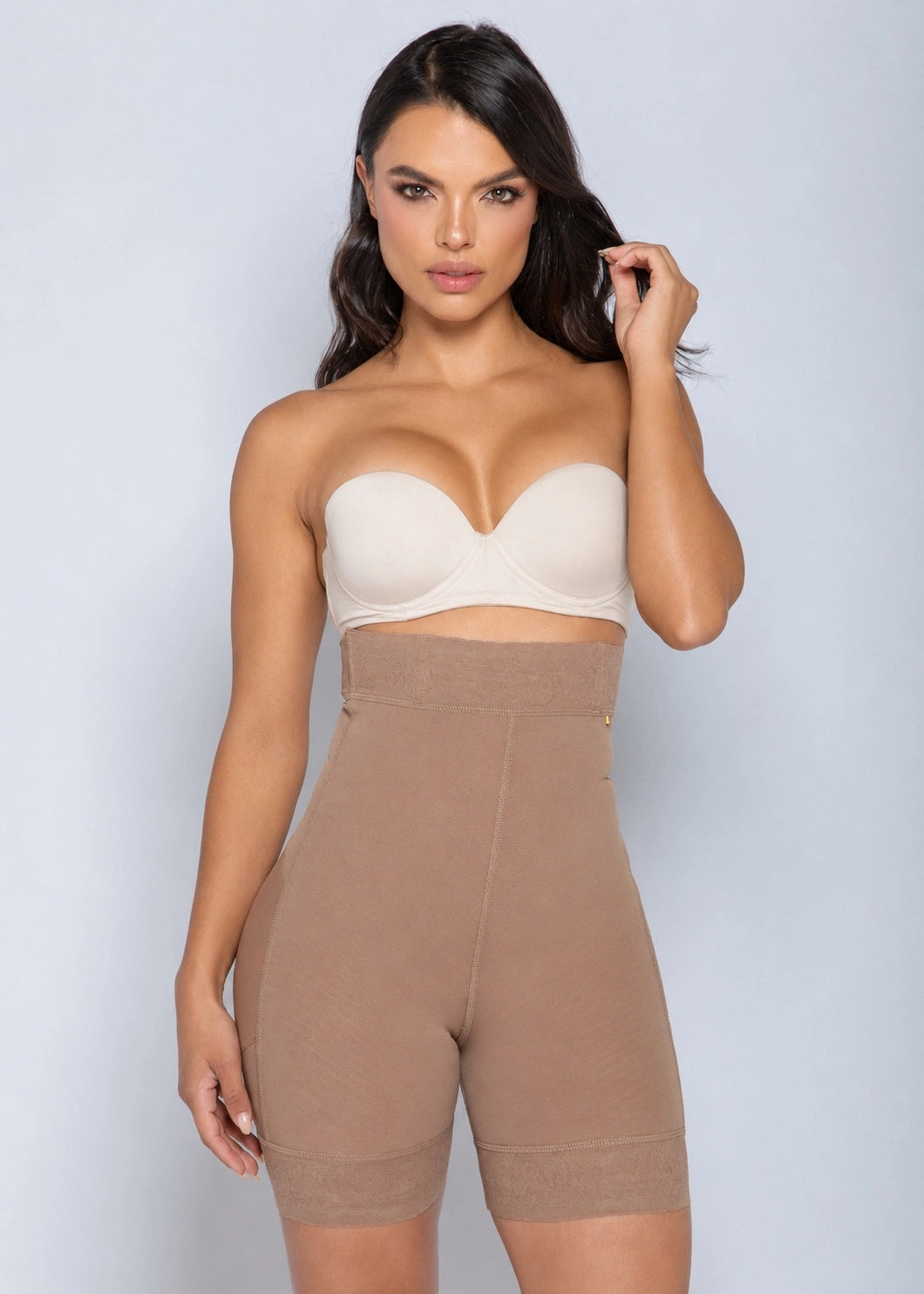 A woman modeling a beige strapless bandeau bra and matching high-waisted shapewear shorts against a light gray studio background.