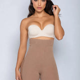 A woman modeling a beige strapless bandeau bra and matching high-waisted shapewear shorts against a light gray studio background.