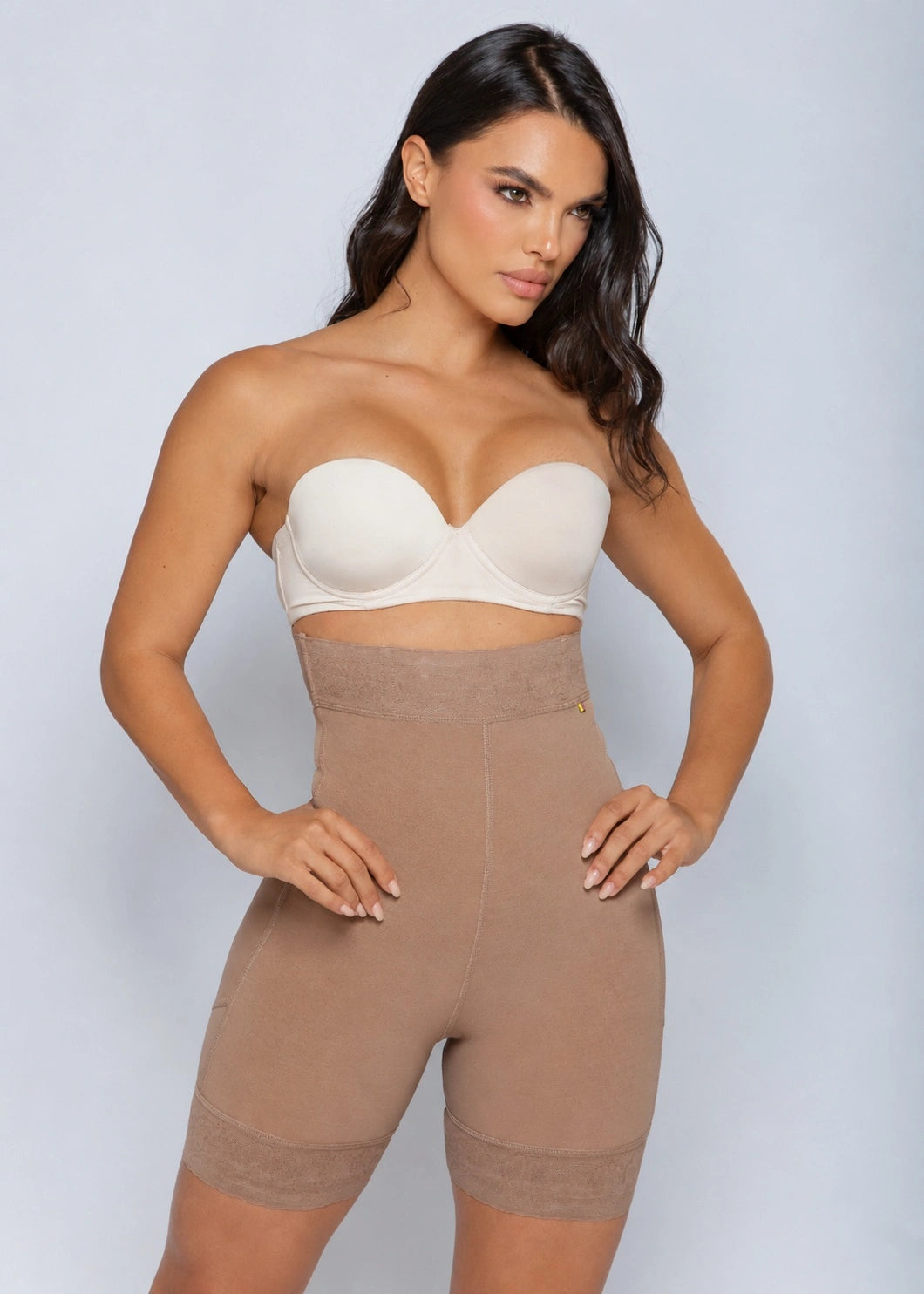 A woman modeling a beige strapless bandeau bra and matching high-waisted shapewear shorts against a light gray studio background.