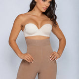 A woman modeling a beige strapless bandeau bra and matching high-waisted shapewear shorts against a light gray studio background.