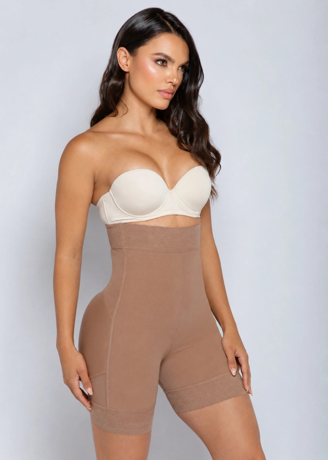 A woman modeling a strapless shapewear bodysuit with a cream underwire bra top and high-waisted tan shorts, set against a plain light gray studio background.
