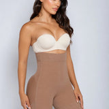 A woman modeling a strapless shapewear bodysuit with a cream underwire bra top and high-waisted tan shorts, set against a plain light gray studio background.