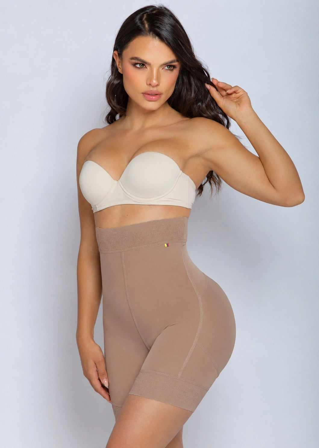 A woman modeling a beige high-waisted shapewear brief and a matching strapless bandeau bra against a light gray studio background.