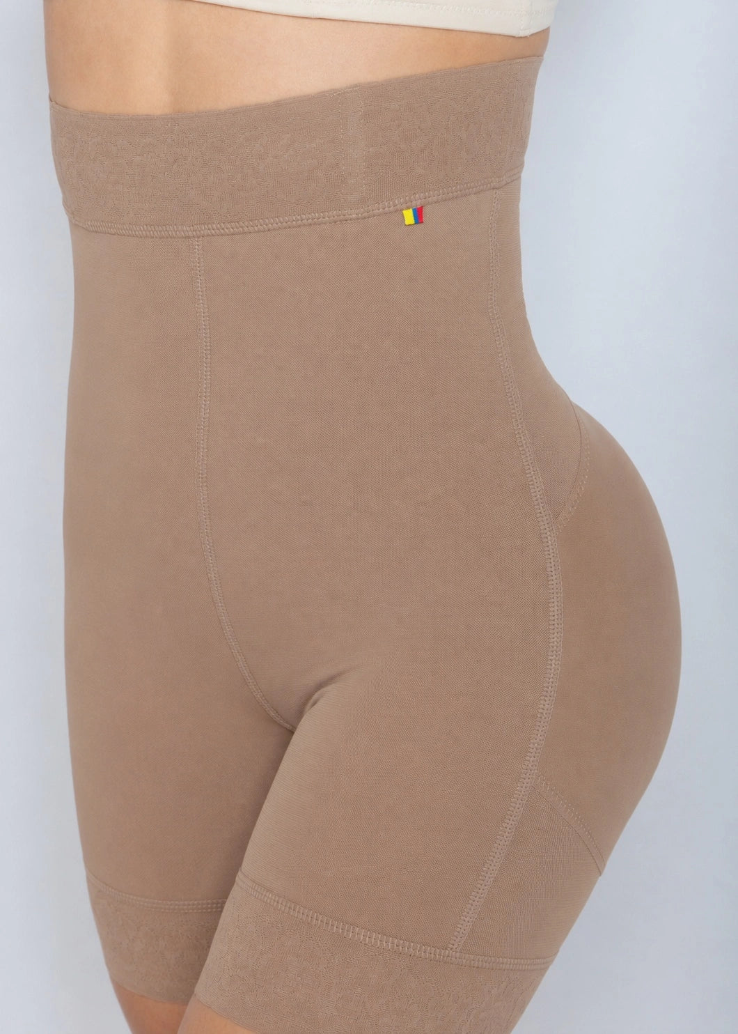 Close-up side view of a person wearing high-waisted beige compression shorts or shapewear against a light gray background.
