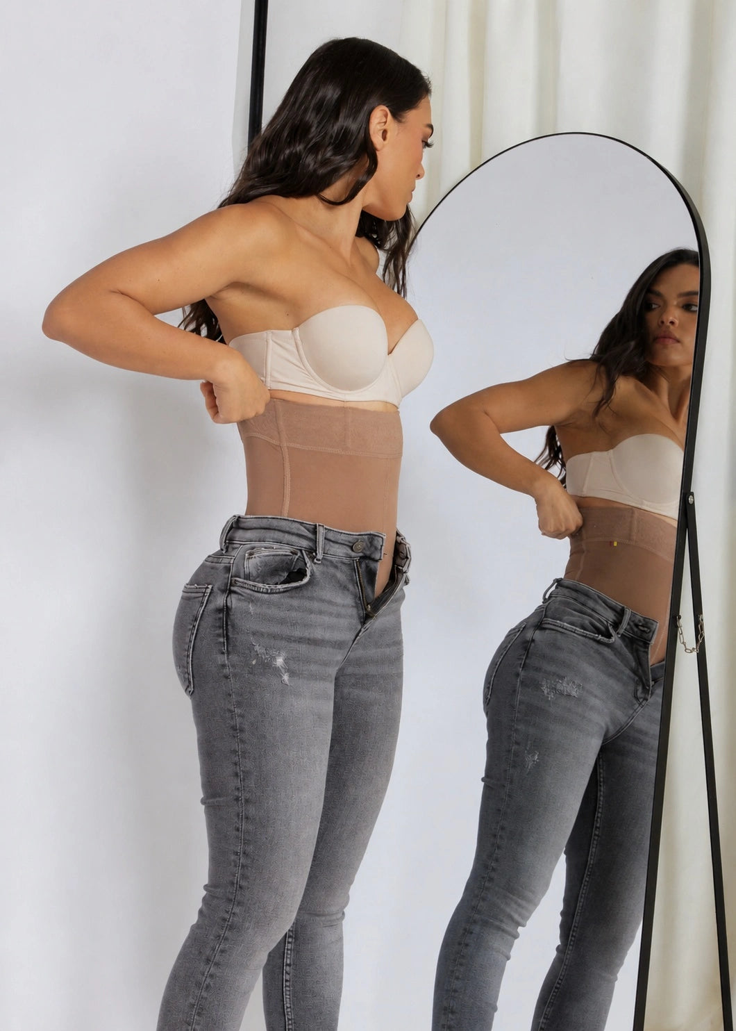 A woman wearing a beige underwire bra and a sheer beige waist trainer shaper, standing in front of a mirror that reflects her side profile. She is wearing gray jeans.