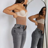 A woman wearing a beige underwire bra and a sheer beige waist trainer shaper, standing in front of a mirror that reflects her side profile. She is wearing gray jeans.
