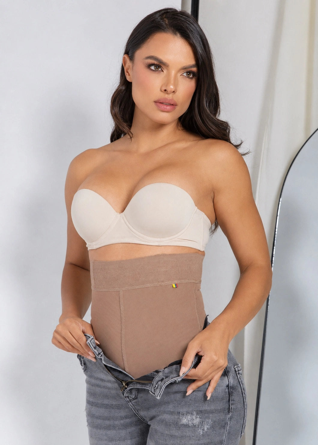 A woman modeling a beige strapless bandeau bra and matching high-waisted shapewear bodysuit, paired with gray jeans.