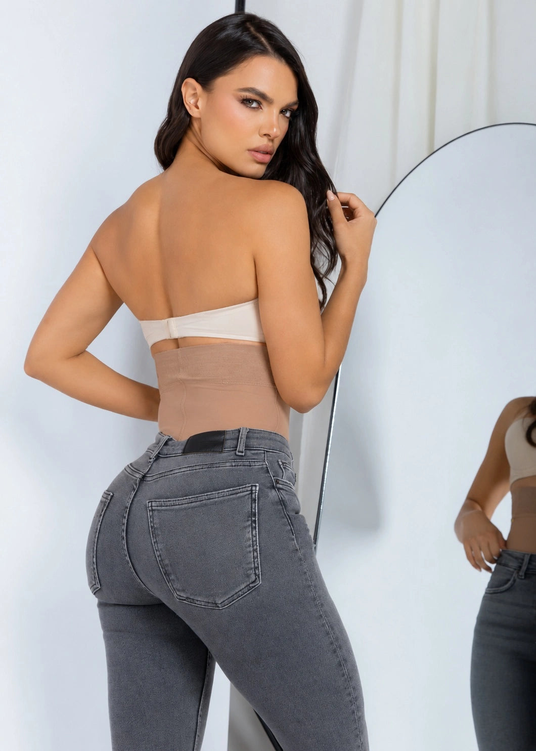 A woman posing from behind wearing a beige strapless bandeau top and gray high-waisted skinny jeans, standing in front of a white studio background with a large oval mirror.