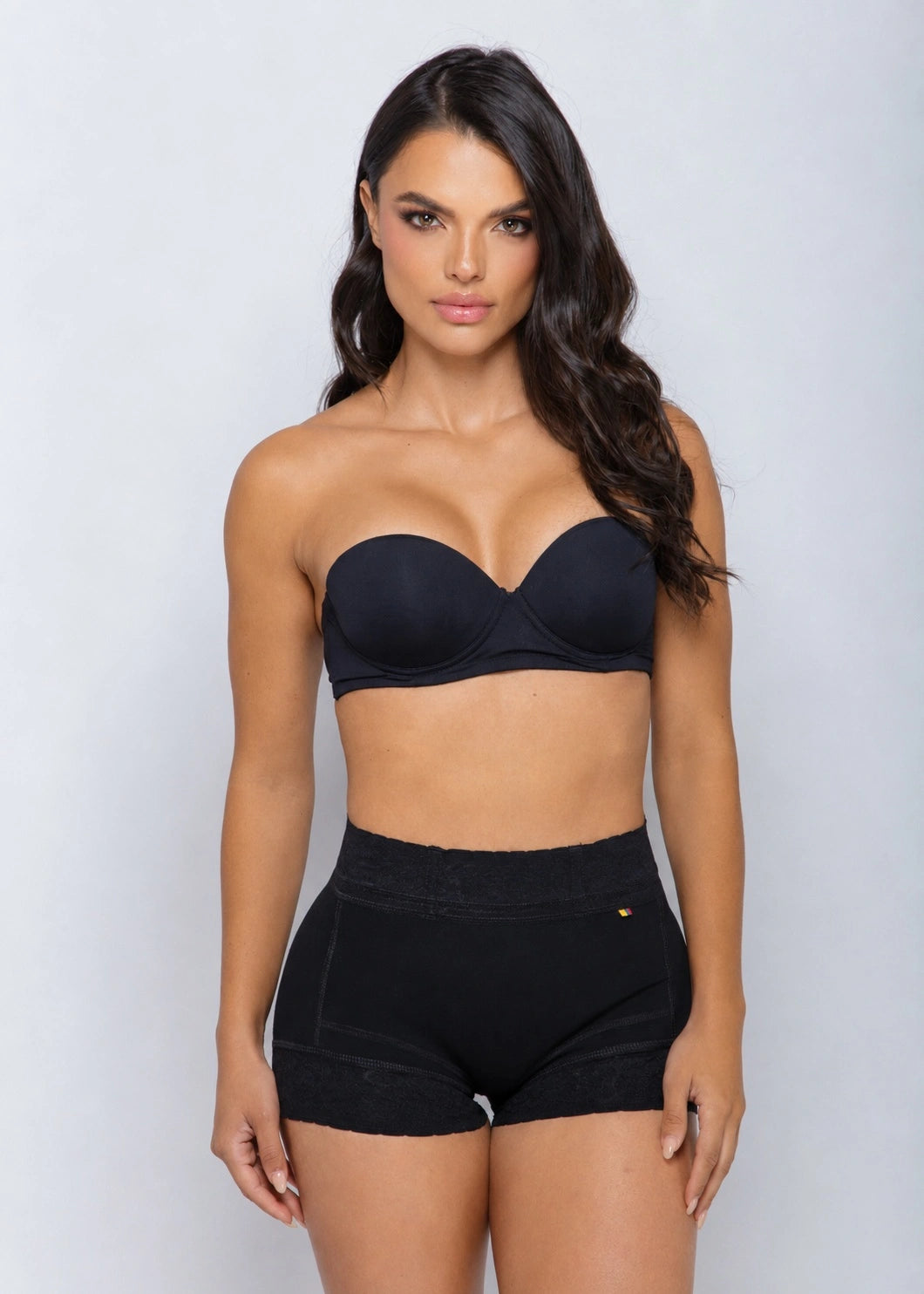 A woman modeling a black strapless bra and matching high-waisted black shapewear shorts against a light gray studio background.