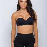 A woman modeling a black strapless bra and matching high-waisted black shapewear shorts against a light gray studio background.