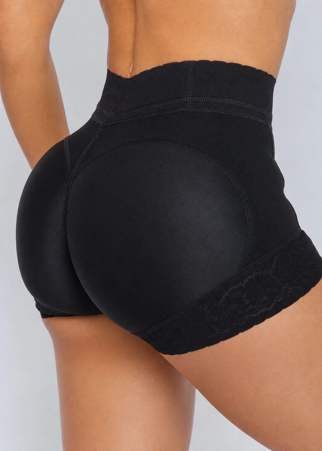 Close-up rear view of a model wearing black high-waisted shapewear shorts with lace trim, shown against a light gray studio background.