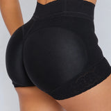 Close-up rear view of a model wearing black high-waisted shapewear shorts with lace trim, shown against a light gray studio background.