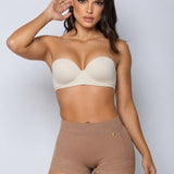 A woman modeling a beige strapless bra and matching high-waisted shapewear shorts against a light gray studio background.