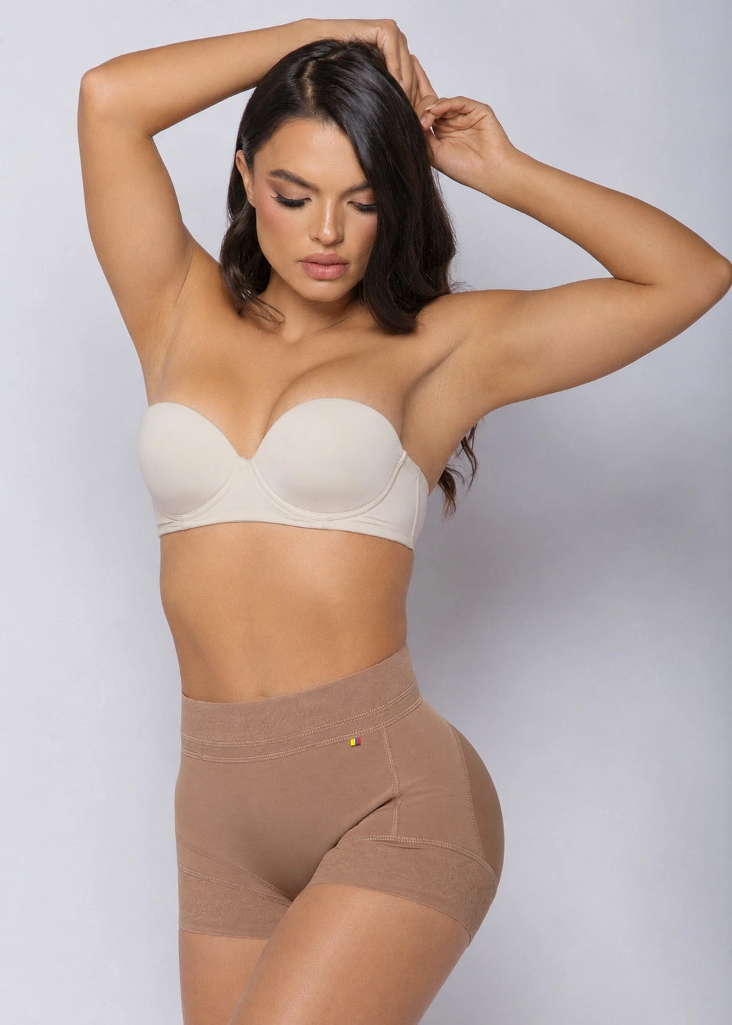 A woman modeling a beige strapless bra and matching high-waisted shapewear briefs against a light gray studio background.