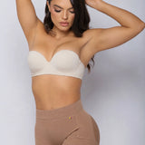 A woman modeling a beige strapless bra and matching high-waisted shapewear briefs against a light gray studio background.