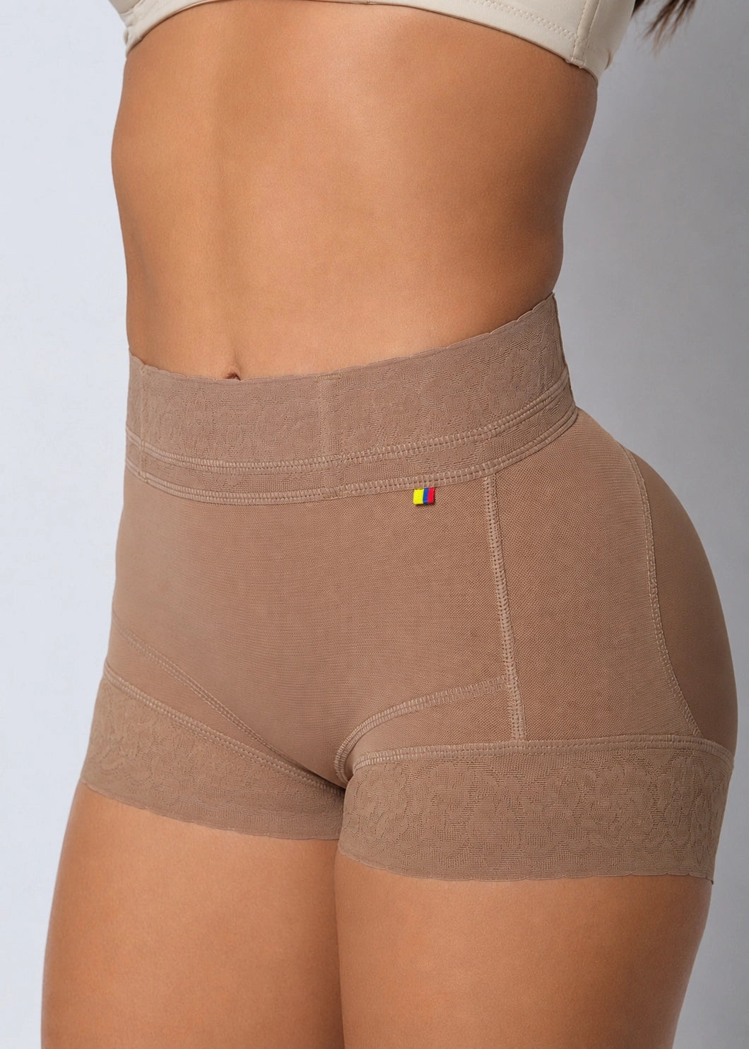 Close-up of a model wearing high-waisted seamless shapewear shorts in a warm taupe-brown color, showing the waistband, hip contours, and leg openings against a neutral gray background.