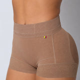 Close-up of a model wearing high-waisted seamless shapewear shorts in a warm taupe-brown color, showing the waistband, hip contours, and leg openings against a neutral gray background.