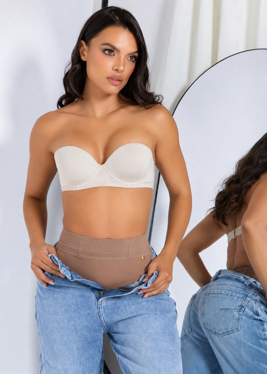 A woman modeling a beige strapless bandeau bra and high-waisted shapewear briefs, with light blue jeans pulled up to show the waist area. A mirror on the right reflects her back and side view.