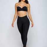 A woman modeling a black strapless bandeau-style bra and matching high-waisted black leggings against a light gray studio background.
