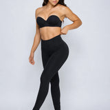 A woman modeling a black two-piece activewear set consisting of a strapless bandeau-style bra and high-waisted leggings, posed against a light gray studio background.