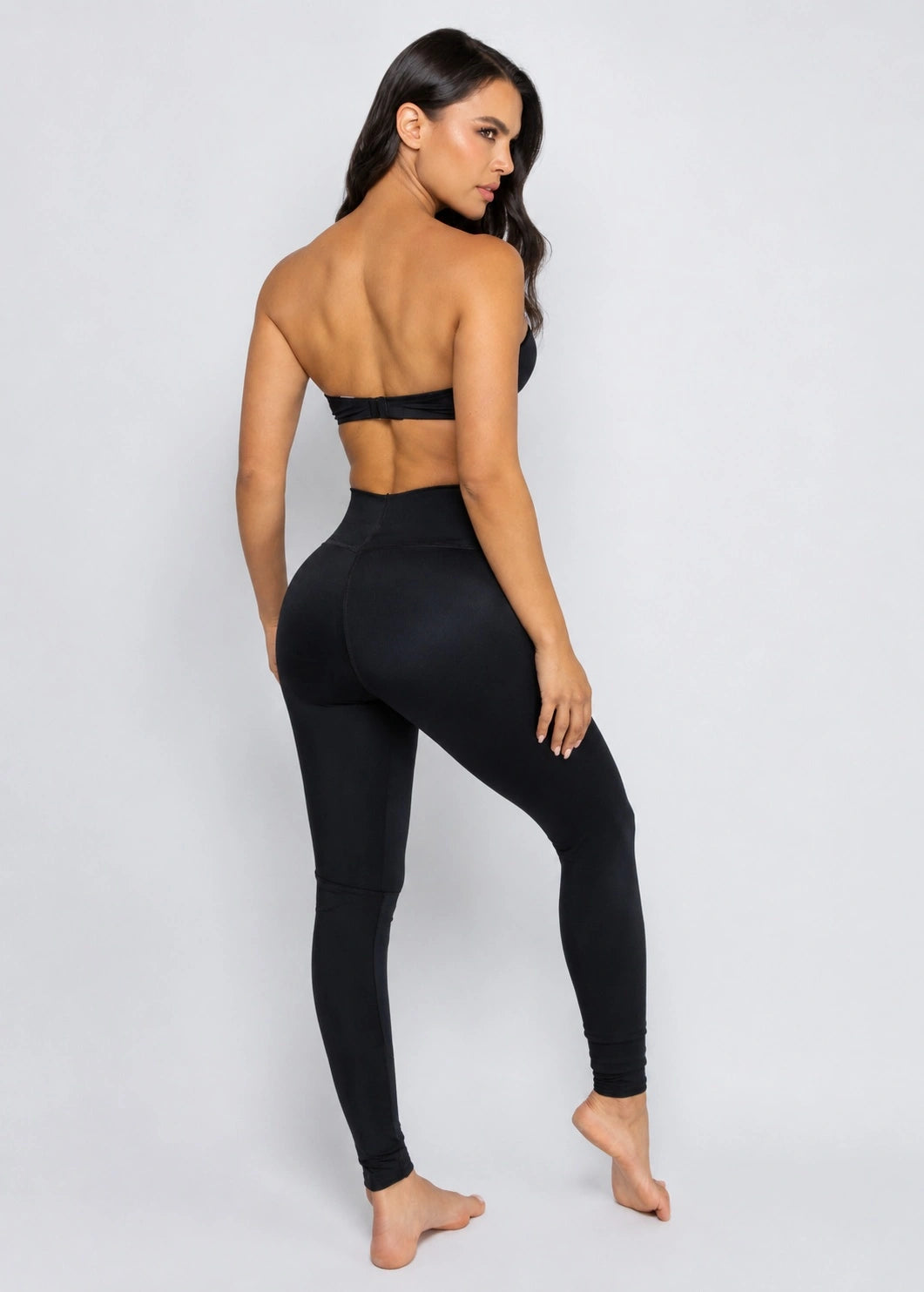 A woman modeling a black two-piece activewear set, featuring a strapless bandeau-style top with a low back cutout and matching high-waisted leggings, shown from a rear three-quarter angle against a light gray studio background.
