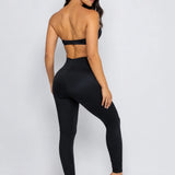 A woman modeling a black two-piece activewear set, featuring a strapless bandeau-style top with a low back cutout and matching high-waisted leggings, shown from a rear three-quarter angle against a light gray studio background.