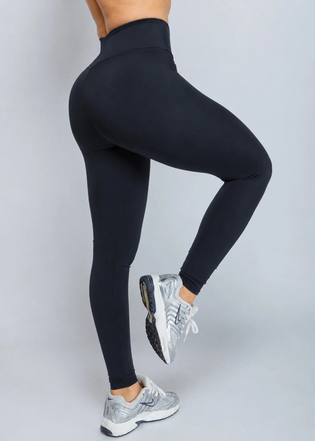 Black high-waisted leggings worn by a model, shown from the side and back as she lifts one leg, paired with silver and white sneakers against a solid gray studio background.