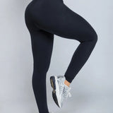 Black high-waisted leggings worn by a model, shown from the side and back as she lifts one leg, paired with silver and white sneakers against a solid gray studio background.
