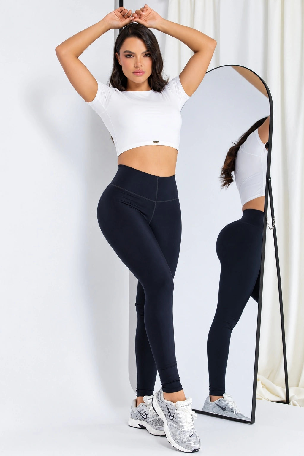 A woman modeling a white short-sleeve crop top and black high-waisted leggings, standing in front of a full-length mirror that shows the back view of the outfit. She is wearing white and silver sneakers against a white studio background with a curtain.