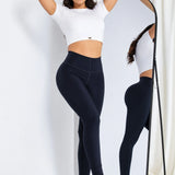 A woman modeling a white short-sleeve crop top and black high-waisted leggings, standing in front of a full-length mirror that shows the back view of the outfit. She is wearing white and silver sneakers against a white studio background with a curtain.