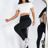 A woman modeling a white short-sleeve crop top and black high-waisted leggings, standing next to a tall floor mirror in a bright studio setting.