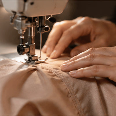 Close-up of a person's hands guiding fabric under a sewing machine needle, stitching a tan garment or material.